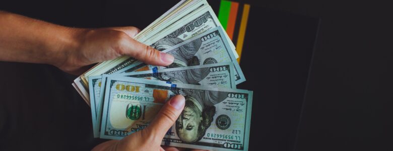 A pair of hands holding a stack of U.S. hundred-dollar bills, fanned out against a dark background with colorful stripes partially visible.