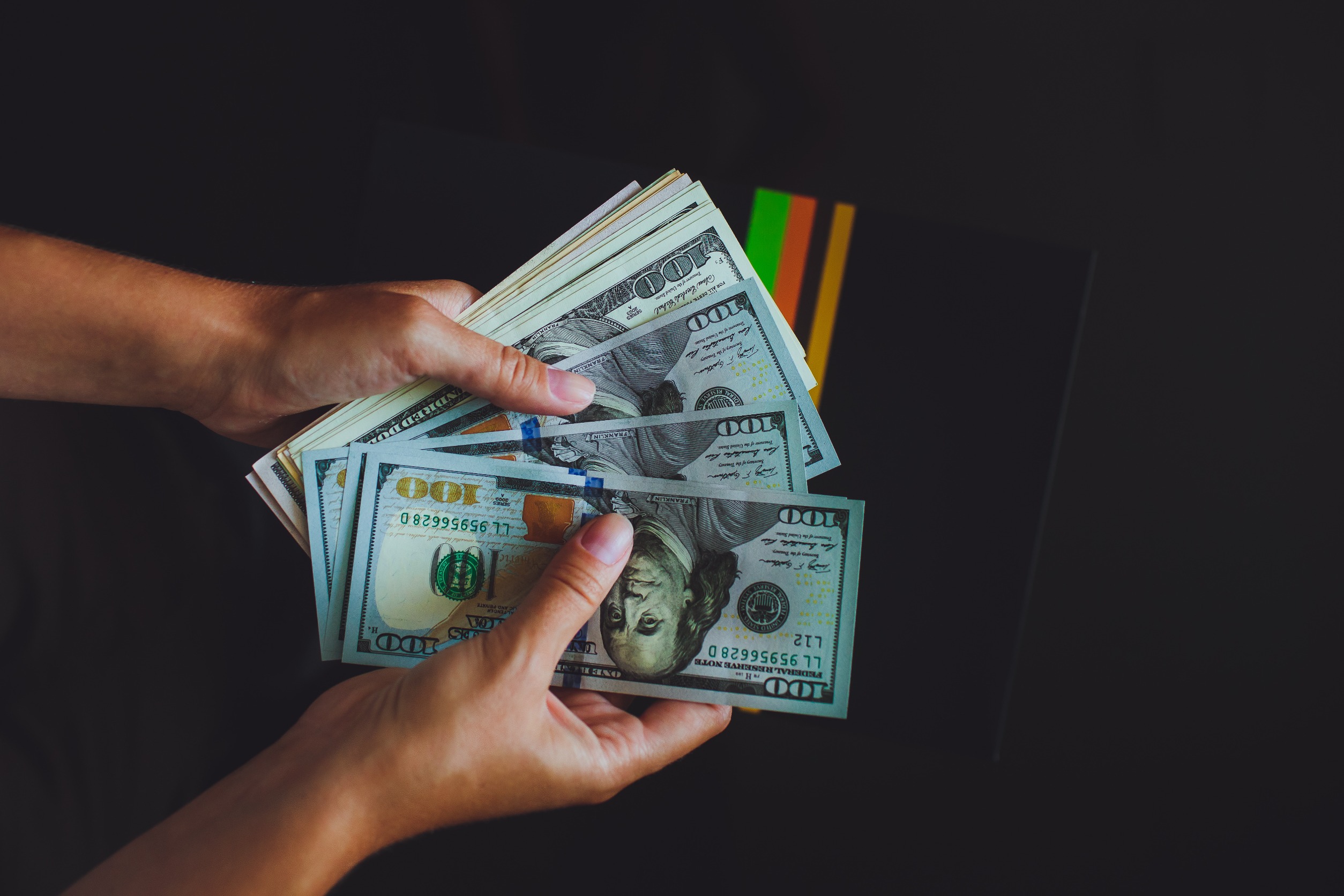 A pair of hands holding a stack of U.S. hundred-dollar bills, fanned out against a dark background with colorful stripes partially visible.