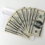 A white envelope partially open on a white surface, with multiple U.S. hundred-dollar bills fanned out in front of it.