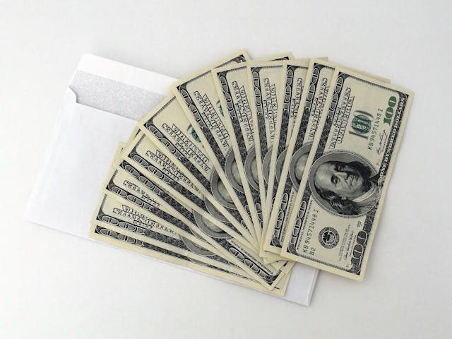 A white envelope partially open on a white surface, with multiple U.S. hundred-dollar bills fanned out in front of it.