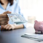 A person in a light blue shirt holding out U.S. hundred-dollar bills, with a pink piggy bank and a stack of additional bills on a dark table in the foreground, against a blurred bright background.