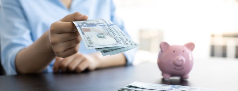 A person in a light blue shirt holding out U.S. hundred-dollar bills, with a pink piggy bank and a stack of additional bills on a dark table in the foreground, against a blurred bright background.