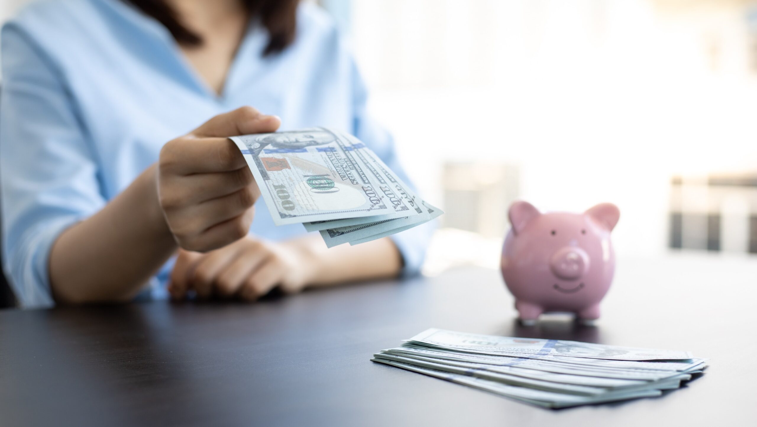 A person in a light blue shirt holding out U.S. hundred-dollar bills, with a pink piggy bank and a stack of additional bills on a dark table in the foreground, against a blurred bright background.