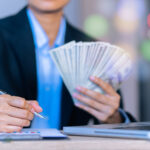 A person in a formal business suit holding a fanned-out stack of cash in one hand while writing with a pen on a document near a laptop, with a blurred background of colorful lights. This could be hard money loans.