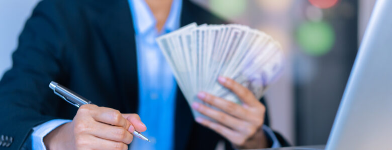 A person in a formal business suit holding a fanned-out stack of cash in one hand while writing with a pen on a document near a laptop, with a blurred background of colorful lights. This could be hard money loans.