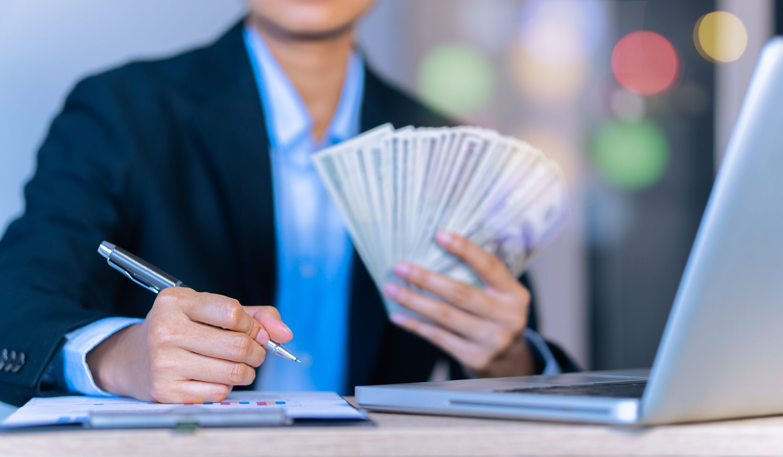 A person in a formal business suit holding a fanned-out stack of cash in one hand while writing with a pen on a document near a laptop, with a blurred background of colorful lights. This could be hard money loans.