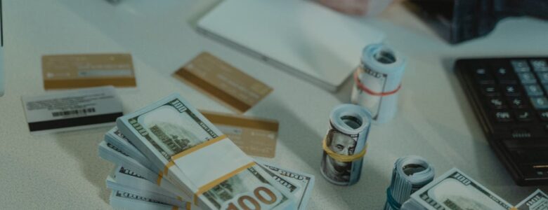 A close-up of a financial workspace featuring stacks and rolls of U.S. hundred-dollar bills, multiple credit cards, a calculator, a notepad, and a person holding a card. The scene suggests financial planning, banking, wealth management or hard money loans.