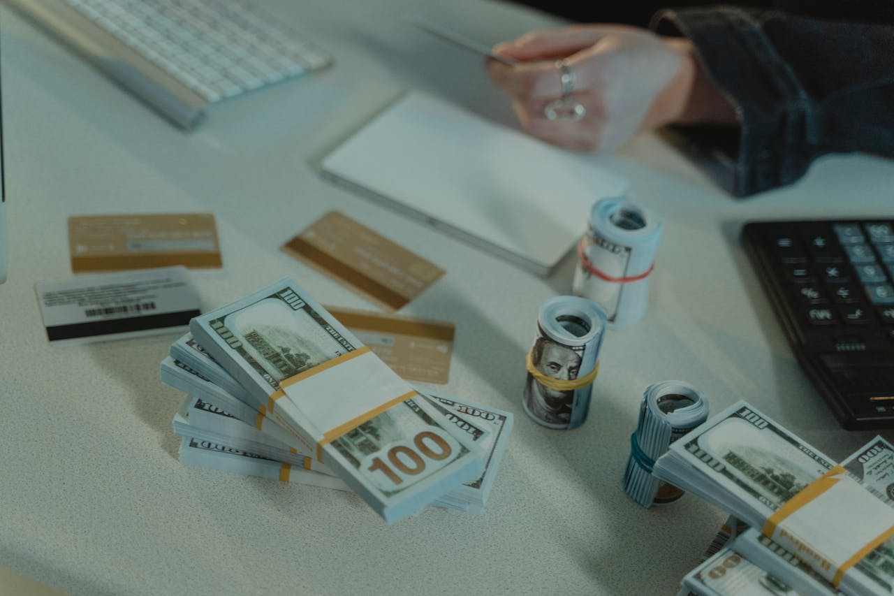 A close-up of a financial workspace featuring stacks and rolls of U.S. hundred-dollar bills, multiple credit cards, a calculator, a notepad, and a person holding a card. The scene suggests financial planning, banking, wealth management or hard money loans.