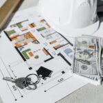 A close-up of real estate planning materials on a table, including a detailed floor plan, a white construction helmet, a stack of U.S. hundred-dollar bills, a set of house keys, and a leveling tool. The image represents property investment, home buying, construction planning.