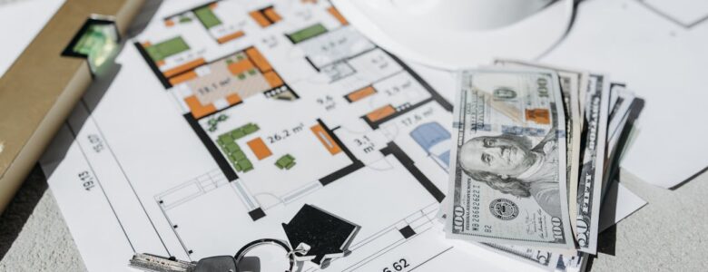 A close-up of real estate planning materials on a table, including a detailed floor plan, a white construction helmet, a stack of U.S. hundred-dollar bills, a set of house keys, and a leveling tool. The image represents property investment, home buying, construction planning.