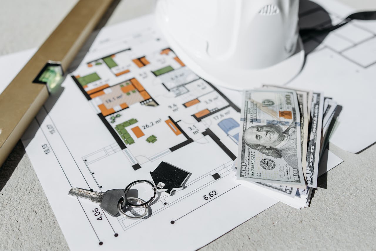 A close-up of real estate planning materials on a table, including a detailed floor plan, a white construction helmet, a stack of U.S. hundred-dollar bills, a set of house keys, and a leveling tool. The image represents property investment, home buying, construction planning.